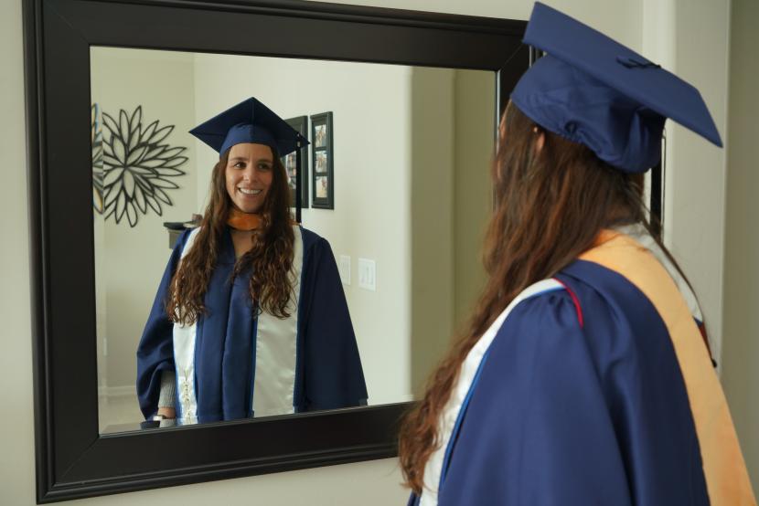 Felicia smiling at her reflection in graduate cap and gown