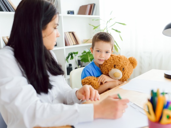 A young boy in a blue shirt holds a large teddy bear while sitting beside a woman in an office setting. The woman, whose face is turned slightly away, appears to be taking notes or drawing. The boy looks directly at the camera with a calm expression.