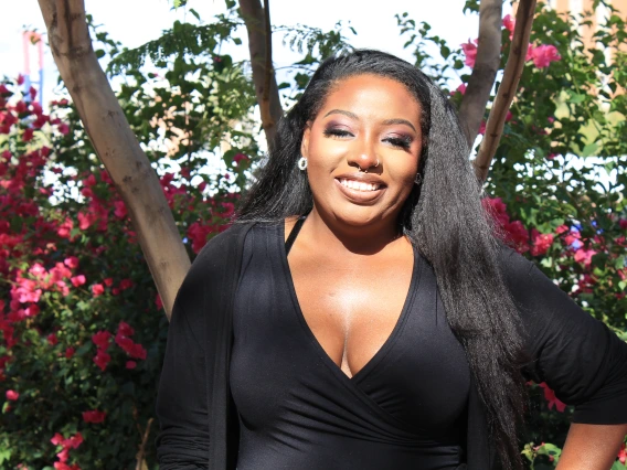  A woman with long black hair and a radiant smile poses outdoors in front of vibrant pink flowers and green foliage. She wears a black V-neck top and a matching cardigan, accessorized with silver hoop earrings and a nose ring. Sunlight highlights her face, creating a warm and confident expression.