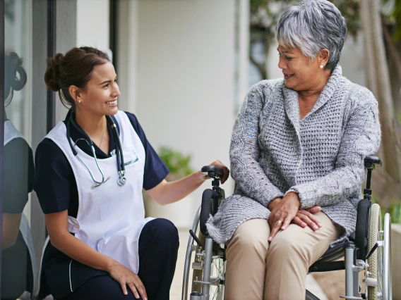 A nurse in a navy-blue uniform and white apron crouches beside an elderly woman seated in a wheelchair. They exchange smiles, conveying warmth and companionship, as they engage in conversation outside a modern building.