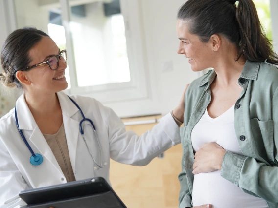 A pregnant woman in a casual green shirt and white top smiles while speaking with her doctor. The doctor, wearing a white coat and glasses, holds a tablet and rests a comforting hand on the patient’s shoulder. The setting is a well-lit clinic