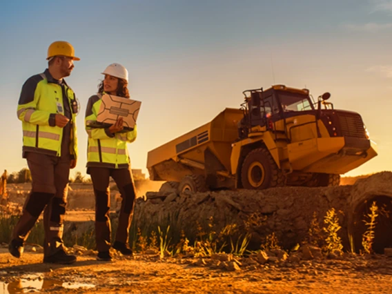 two people in hi-viz vests walking outdoors next to a piece of heavy machinery