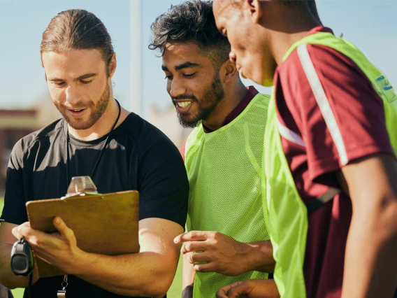 a coach writing on a clipboard, with sports players in neon yellow jerseys huddled around him. 