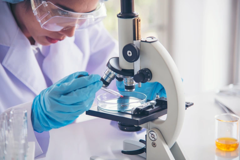 A scientist wearing protective goggles and blue gloves uses a pipette to transfer a red liquid into a petri dish under a microscope, conducting laboratory research in a bright, clinical setting.