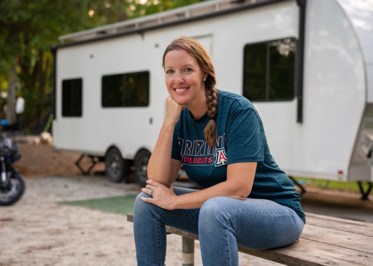 Image of Angela Kirby sitting on a wooden picnic table in front of her RV. 
