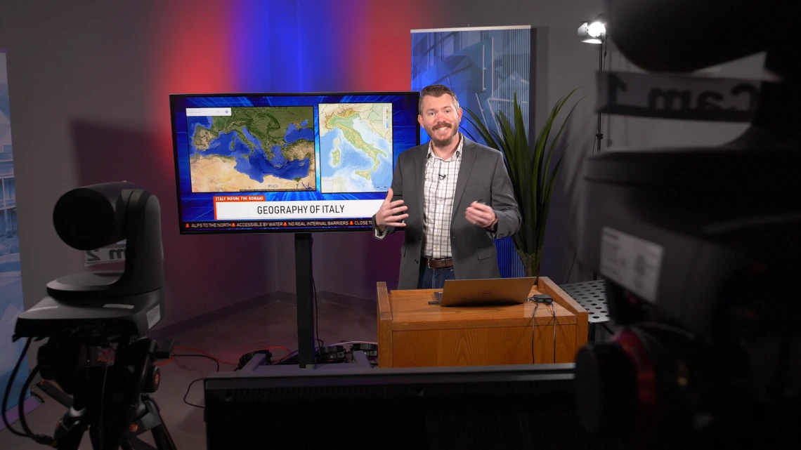 A male professor of geography teaches to multiple video cameras while gesturing to a large display screen behind him