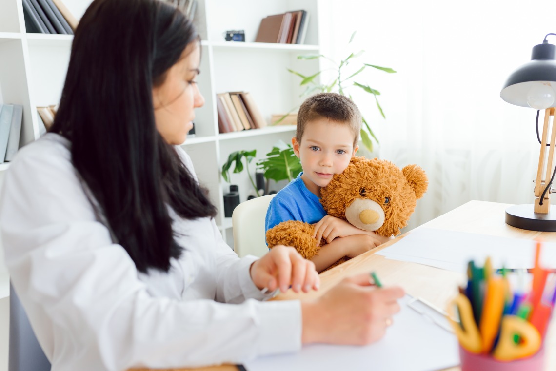 A young boy in a blue shirt holds a large teddy bear while sitting beside a woman in an office setting. The woman, whose face is turned slightly away, appears to be taking notes or drawing. The boy looks directly at the camera with a calm expression.