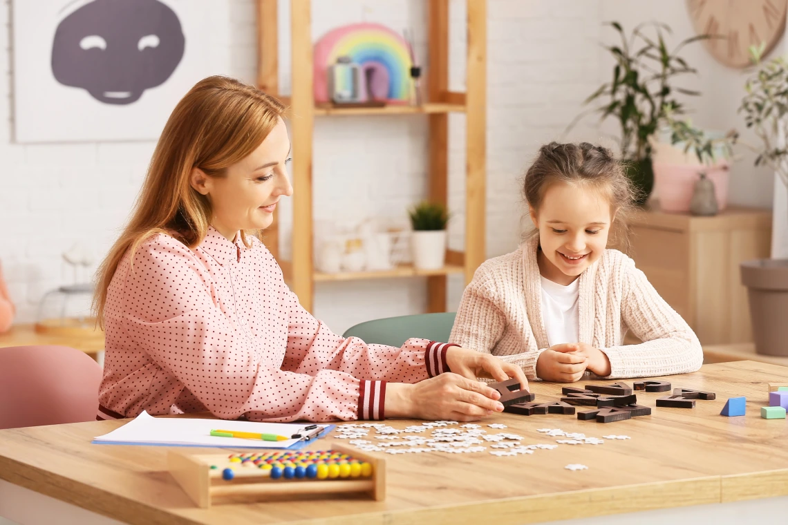 A woman and a child sit at a table filled with puzzle pieces and colorful educational tools. The woman guides the child as they focus on building letters, sharing smiles in a warm, organized space.