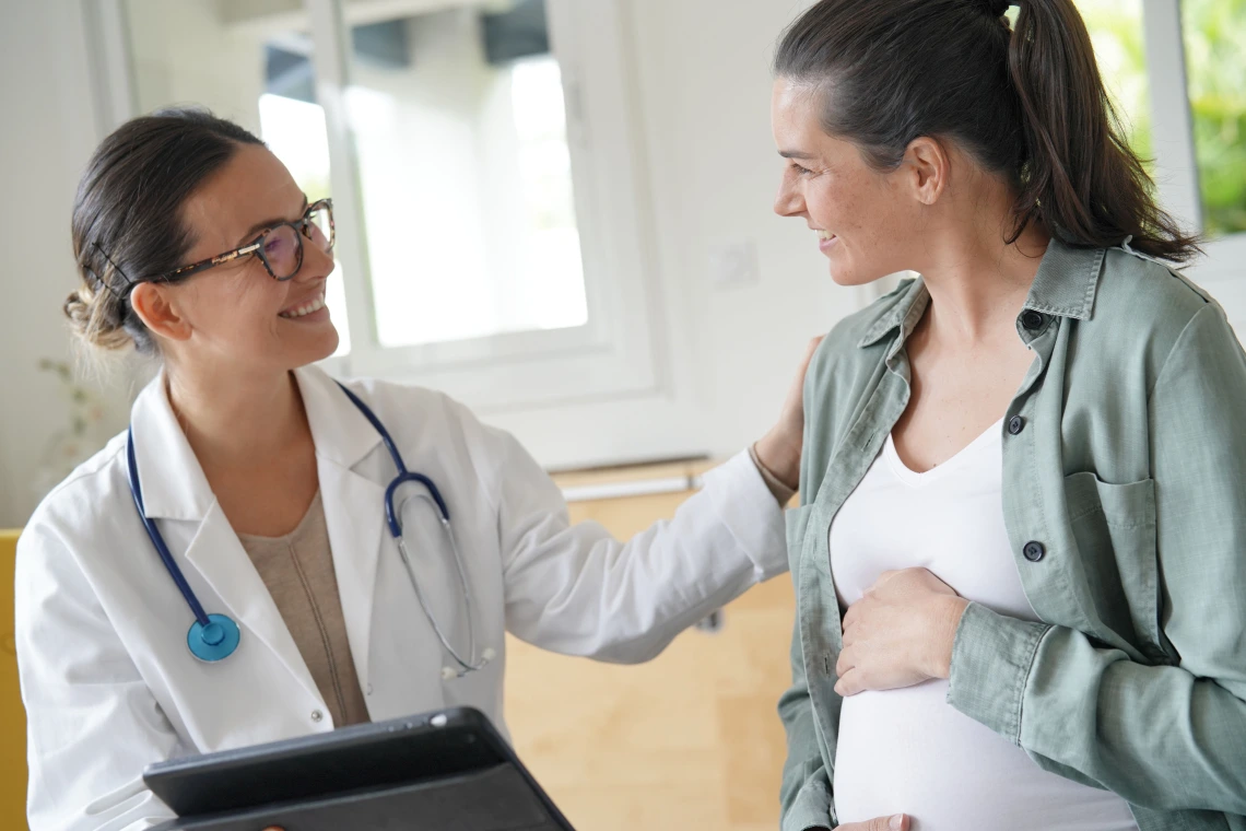 A pregnant woman in a casual green shirt and white top smiles while speaking with her doctor. The doctor, wearing a white coat and glasses, holds a tablet and rests a comforting hand on the patient’s shoulder. The setting is a well-lit clinic