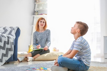 An adult and child sit on the floor. The adult is observing the child as he gazes up at the ceiling.