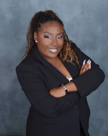 A woman with a warm smile poses confidently with her arms crossed against a textured gray backdrop. She wears a black blazer over a white top, accessorized with a black wristwatch, a bracelet, and stud earrings. Her hair is styled in a curly half-up, half-down look, and her makeup features soft, neutral tones.