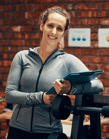 A smiling woman in athletic wear posing with a clipboard in a fitness studio.