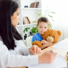 A young boy in a blue shirt holds a large teddy bear while sitting beside a woman in an office setting. The woman, whose face is turned slightly away, appears to be taking notes or drawing. The boy looks directly at the camera with a calm expression.