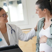 A pregnant woman in a casual green shirt and white top smiles while speaking with her doctor. The doctor, wearing a white coat and glasses, holds a tablet and rests a comforting hand on the patient’s shoulder. The setting is a well-lit clinic