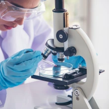 A scientist wearing protective goggles and blue gloves uses a pipette to transfer a red liquid into a petri dish under a microscope, conducting laboratory research in a bright, clinical setting.