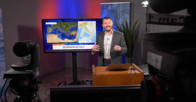 A male professor of geography teaches to multiple video cameras while gesturing to a large display screen behind him