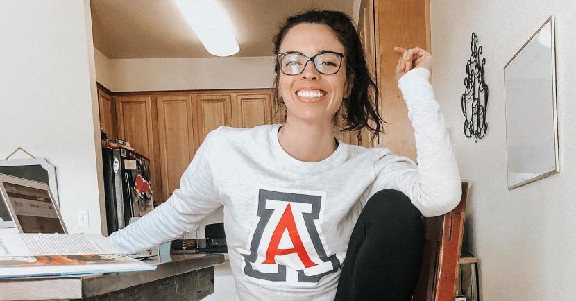 Kellie Kirsch sitting at a table with laptop in UArizona A Shirt