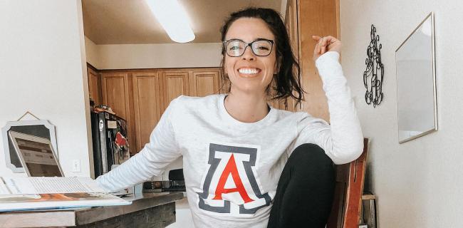 Kellie Kirsch sitting at a table with laptop in UArizona A Shirt